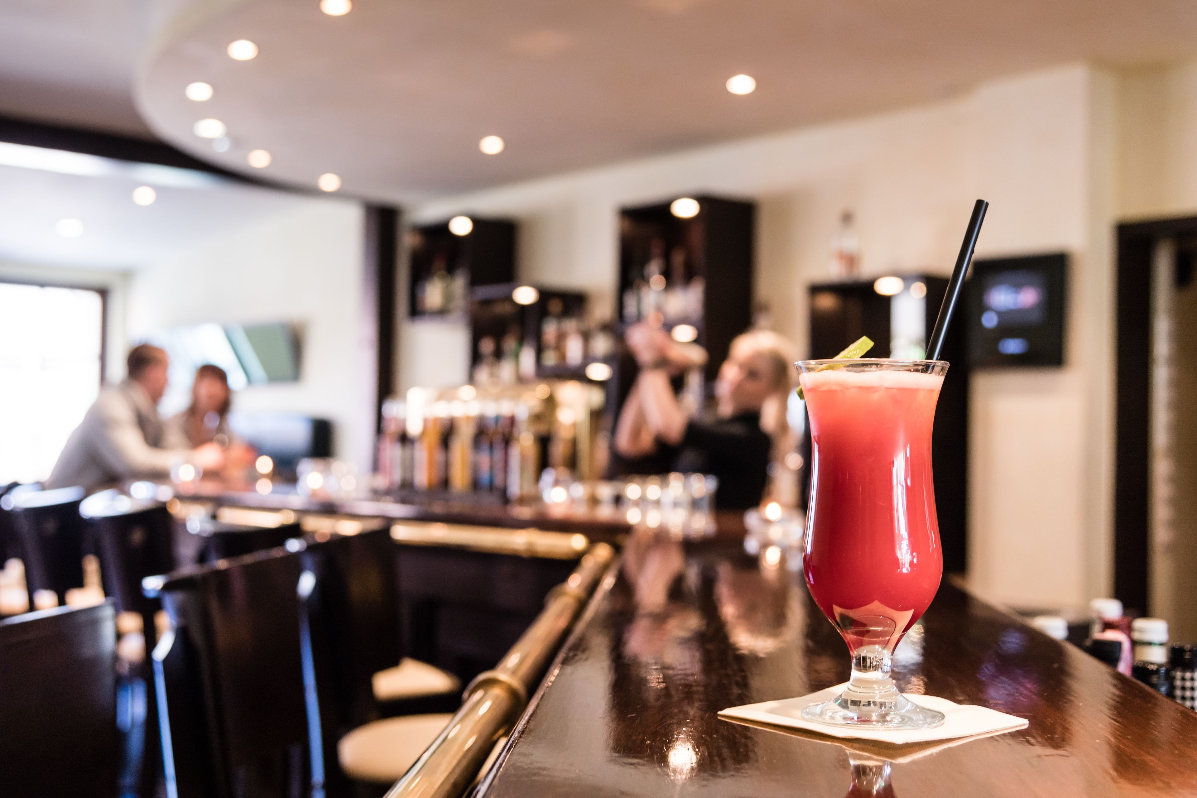 Eine Bar mit einem Glas rotem Getränk auf der Theke, Barhocker und einem Fernsehbild im Hintergrund.