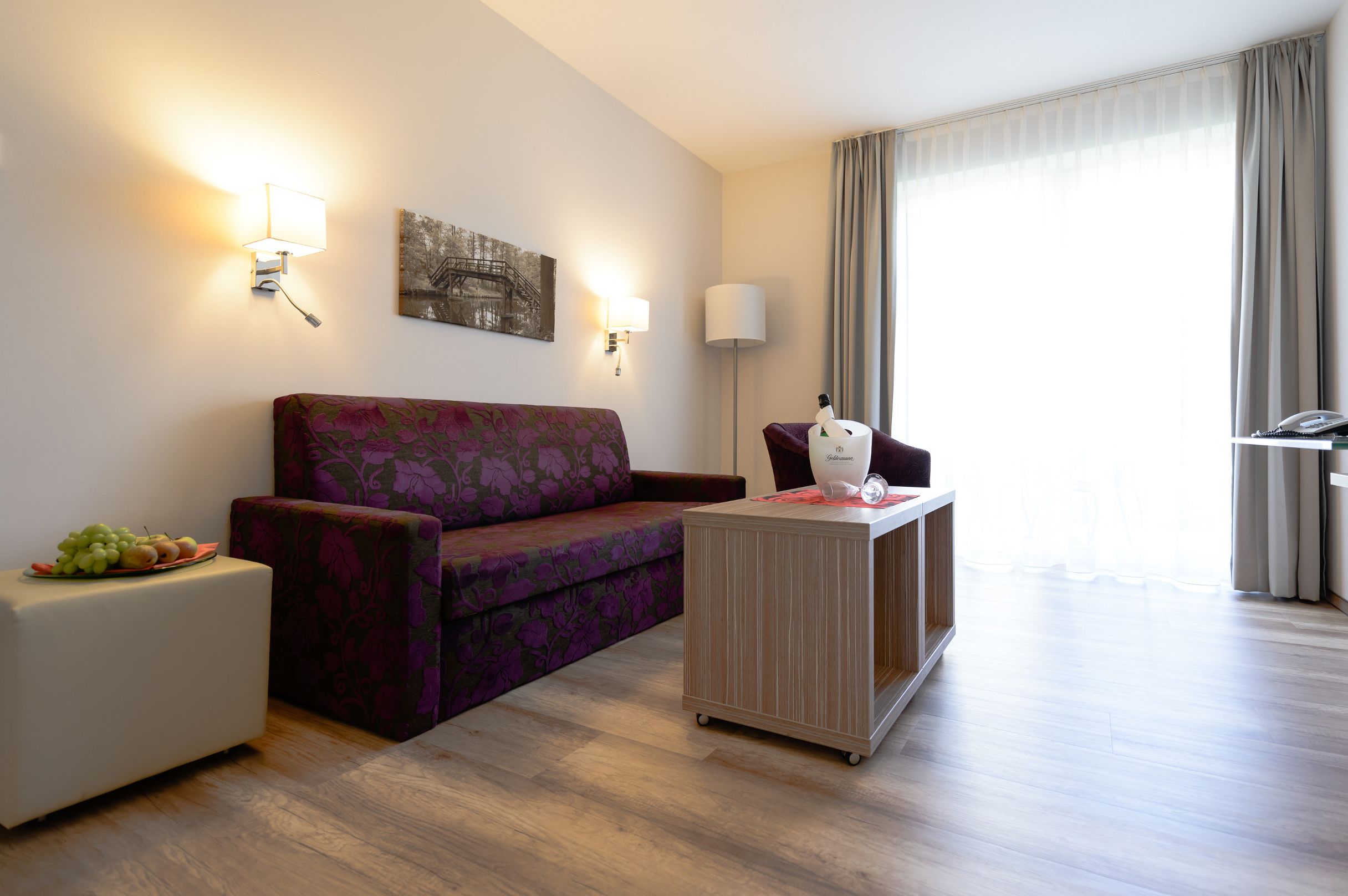 A modern hotel room with a purple sofa, a small side table with fruit, a lamp, and a painting on the wall.