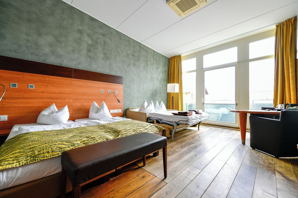 A hotel room with a large bed, a smaller bed, a desk, and a view of the city through large windows.