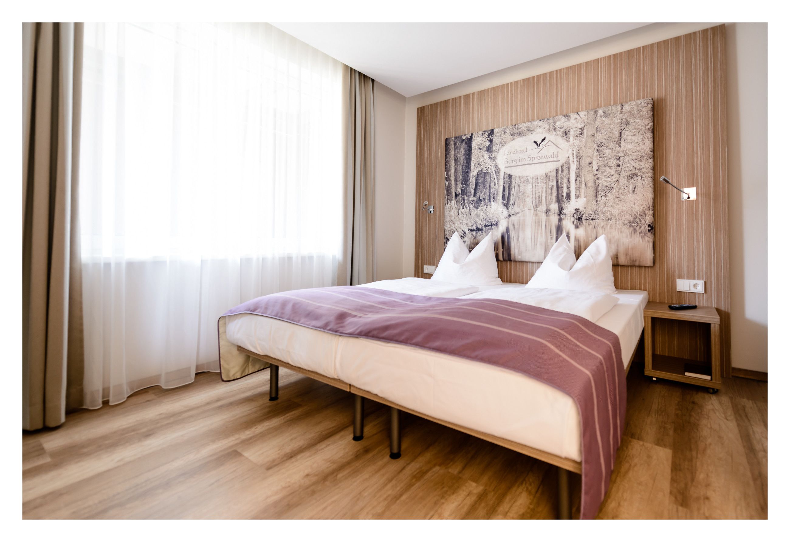 Une chambre moderne avec un grand lit, un plancher en bois, une fenêtre ornée de rideaux légers et une œuvre d’art accrochée au mur.