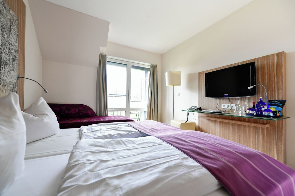 A hotel room with a bed, a TV mounted on the wall, a desk with a lamp, and a window with curtains.