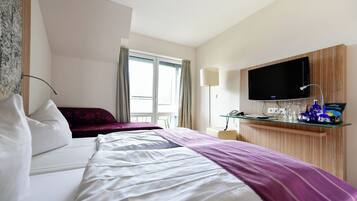 A hotel room with a bed, a TV mounted on the wall, a desk with a lamp, and a window with curtains.