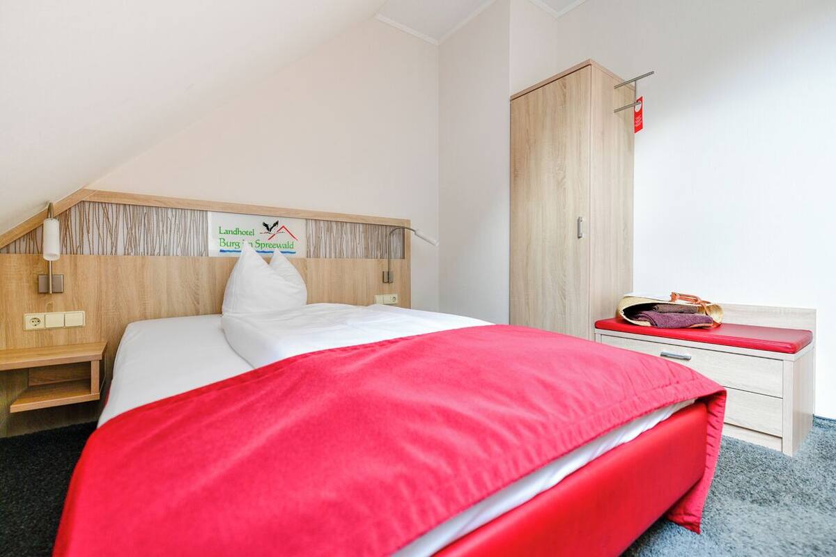 A hotel room with a bed, a wooden headboard, a nightstand, a wardrobe, and a red bench.