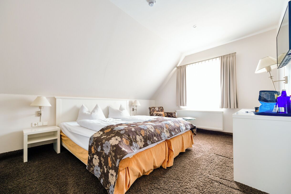 A hotel room with a bed, a desk, a chair, a lamp, and a window with curtains.