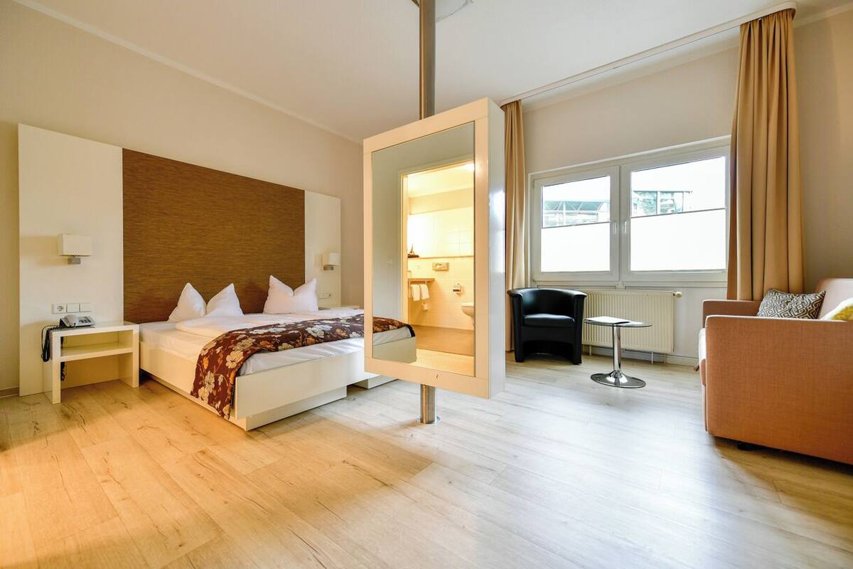 A modern hotel room with a large bed, a sitting area with a sofa and armchair, a small table, and a large mirror.