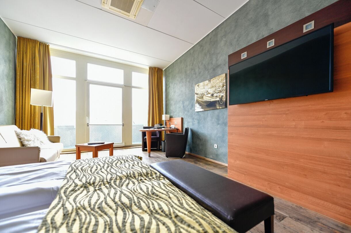 Habitación de hotel con cama, escritorio, silla, banqueta, televisor y ventana con cortinas.