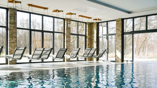 Una piscina interior con paredes de vidrio transparente, múltiples sillones de descanso y vistas a árboles.