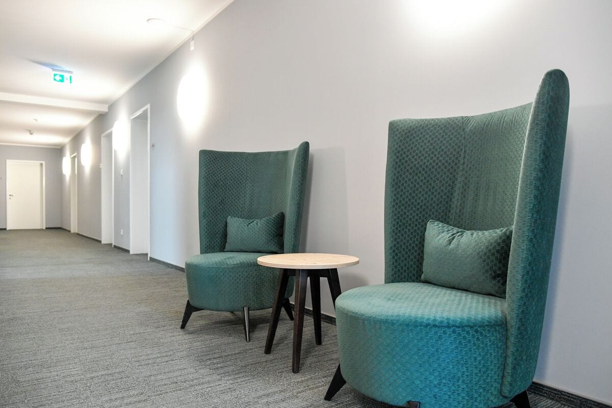 Un couloir avec deux fauteuils turquoise et une petite table ronde, un sol recouvert de moquette et une porte au fond.