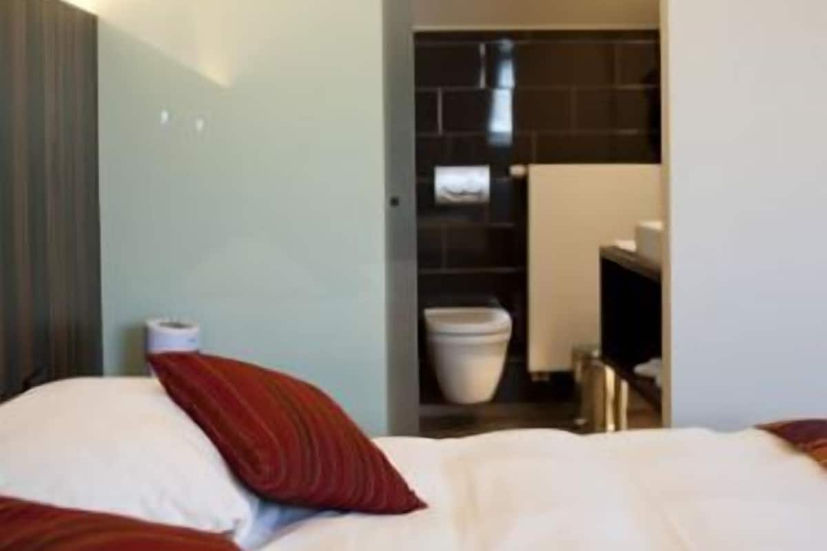 Habitación de hotel con cama, almohadas, mesita de noche y baño con inodoro y lavamanos.