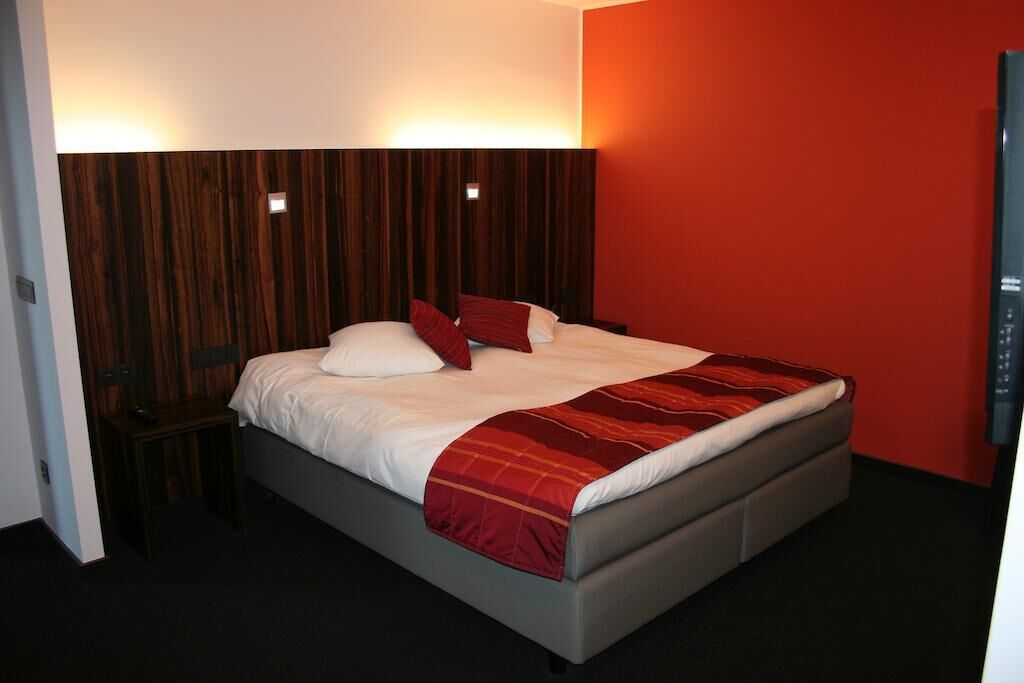 A modern bedroom with a wooden headboard, a bed with white bedding and a red throw blanket, and a nightstand with a lamp.