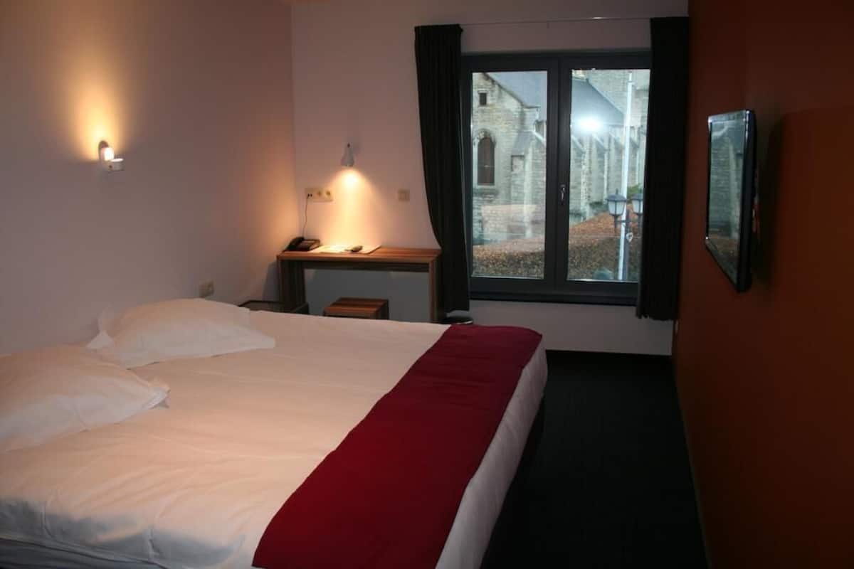 Habitación de hotel con cama, escritorio, silla, ventana con cortinas y una lámpara fijada a la pared.