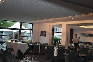 A restaurant with tables set for dining, large windows, and a waiter in the background. - Hotel Carpinus (Herent)
