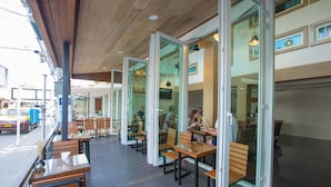 An outdoor café with wooden tables and chairs, glass doors leading to the interior, and a view of the street. - P168 Hostel Samui (Koh Samui)