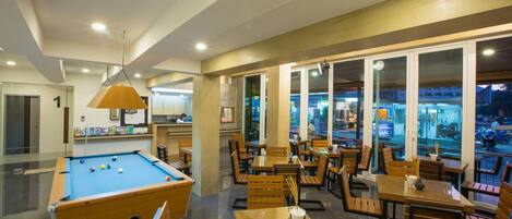 A billiards table in a well-lit room with wooden chairs and tables, large windows, and a view of the outside.