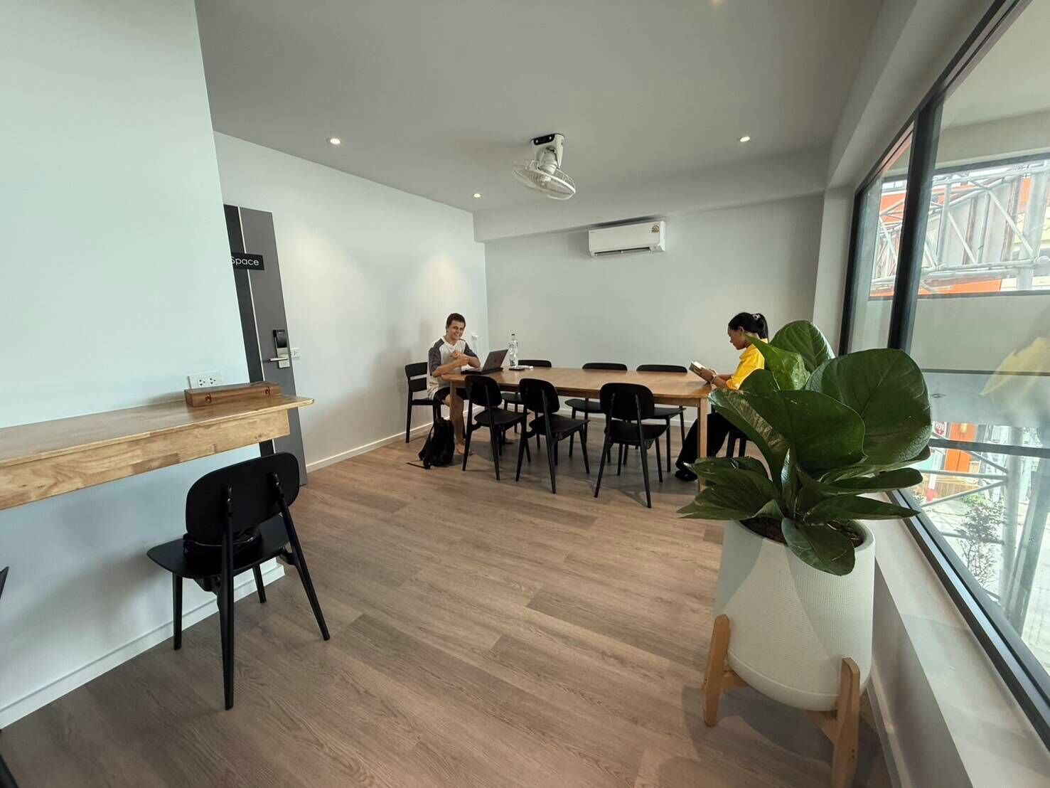 Una sala de reuniones moderna con una larga mesa, sillas negras, un escritorio de madera, una planta en maceta y un equipo de aire acondicionado.