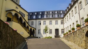 A courtyard with a central building featuring a dark roof and multiple windows, flanked by yellow buildings with arched windows and a stone wall with flowers. - Katholisch-Soziales Institut (Siegburg)