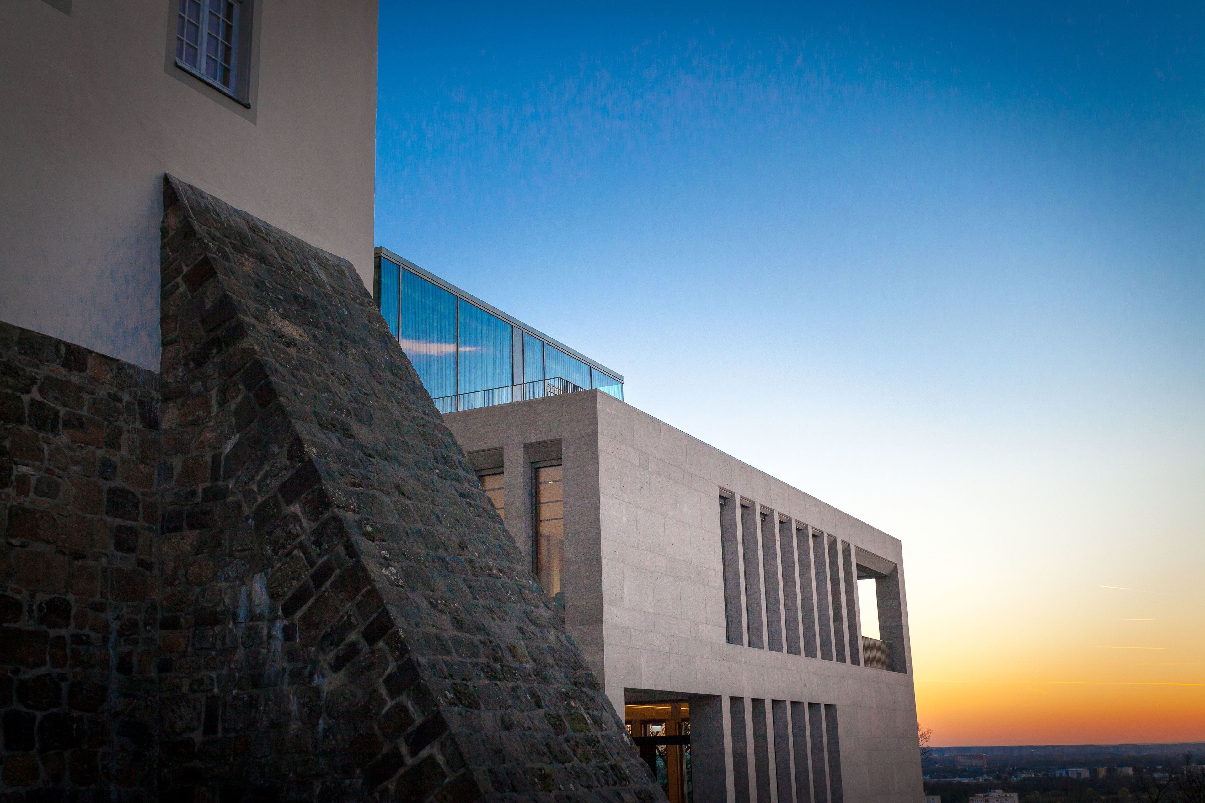 Un bâtiment moderne avec une façade en pierre et un balcon en verre donnant sur une vue panoramique de la ville au coucher du soleil.