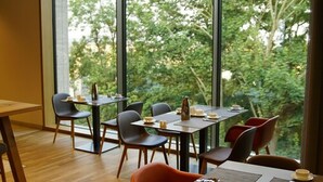 A dining area with tables and chairs, large windows, and a view of trees.