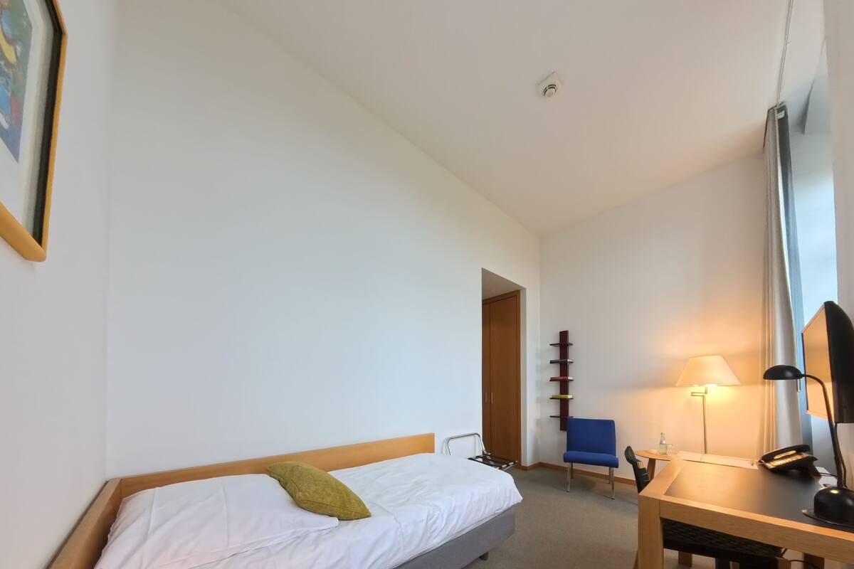 A hotel room with a bed, desk, chair, lamp, and a door leading to another room.