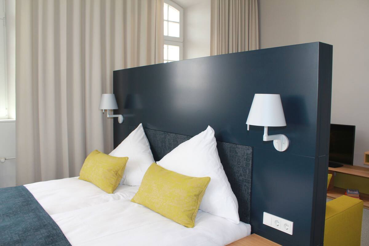 A hotel room with a bed, two bedside lamps, a television, and a window with curtains.