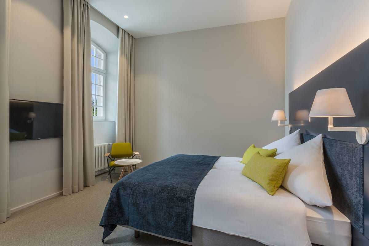 A modern hotel room with a bed, two bedside lamps, a TV, a small table with a chair, and a window with curtains.