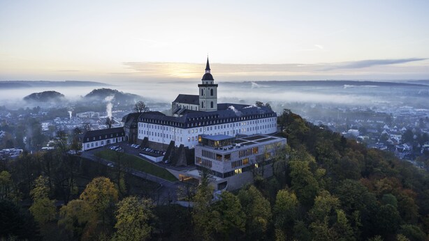 A large, historic building with a prominent tower, surrounded by lush greenery and a foggy landscape.