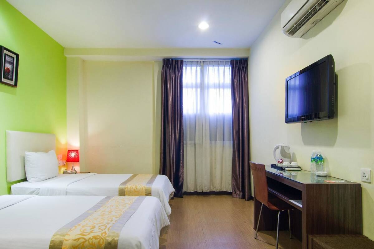 A hotel room with two beds, a desk, a television, and an air conditioning unit.