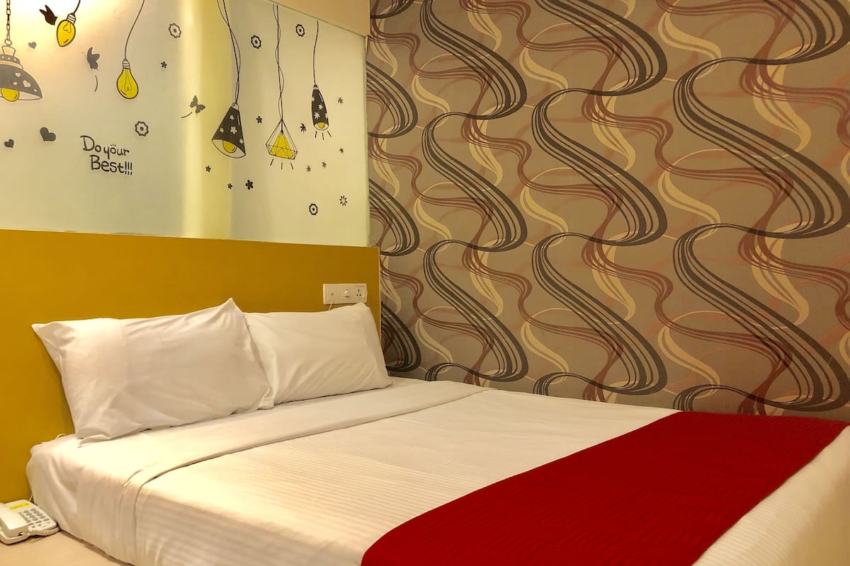 A bed with white bedding and a red throw blanket, a white phone on the side, and a wall with a floral pattern and a light fixture with hanging bulbs.