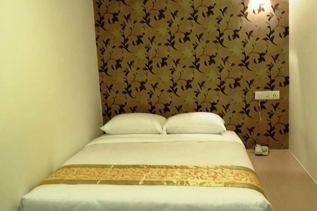 A neatly made bed with white linens and a patterned bedspread, a wall with a floral wallpaper design, and a wall-mounted light fixture.
