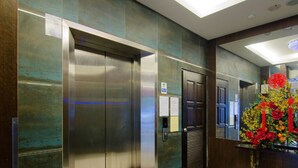 Ein moderner Aufzug mit Metalltüren und digitaler Anzeige, ein Empfangstisch mit Blumenarrangement und eine Wand mit strukturierter Oberfläche.