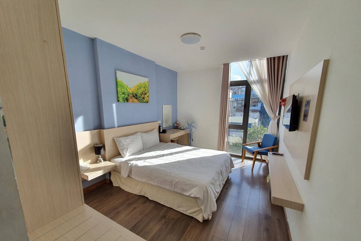 Ein Hotelzimmer mit Bett, Nachttisch, Lampe und Blick auf die Stadt durch ein großes Fenster.