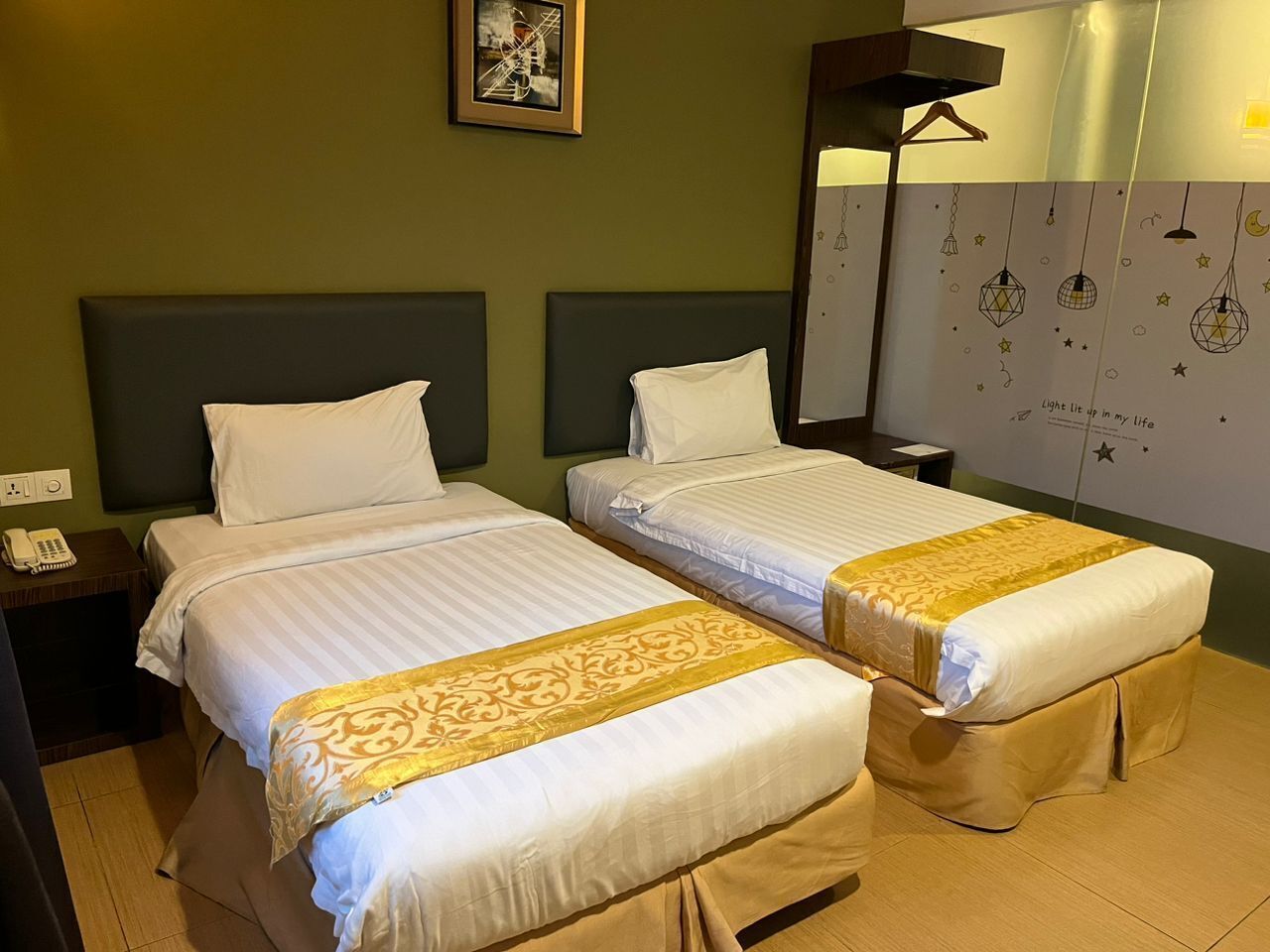 A hotel room with two beds, a bedside table with a phone, a wall-mounted lamp, and a decorative wall with hanging lights.