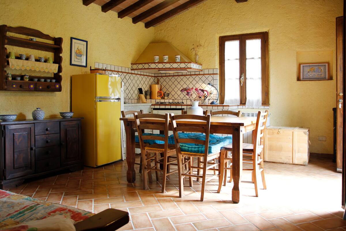 A kitchen with a yellow refrigerator, wooden chairs, a tiled floor, and a window with wooden shutters.