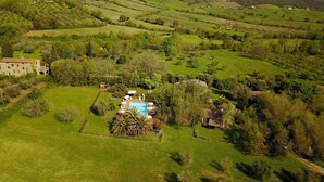 A rural landscape with rolling hills, a large stone building, and a swimming pool surrounded by greenery. - Casale Vigna Carbona (Gavorrano)