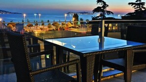 An outdoor seating area with a glass table and wicker chairs, overlooking a coastal view with street lights and a distant cityscape. - Dragos Island Hotel (Istanbul)