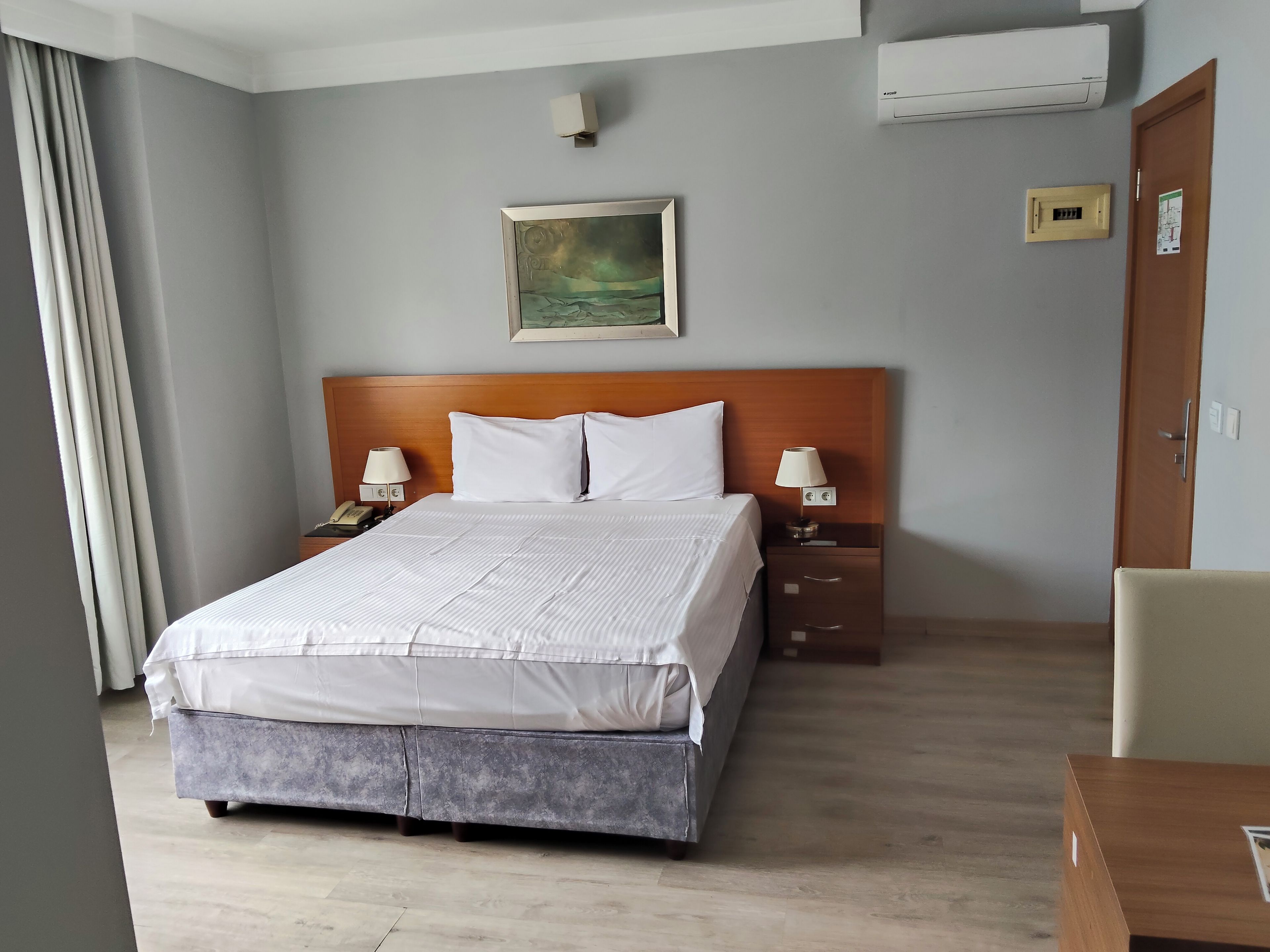 A hotel room with a large bed, two bedside tables with lamps, a wooden headboard, a painting on the wall, an air conditioning unit, a door with a handle, and a wooden floor.