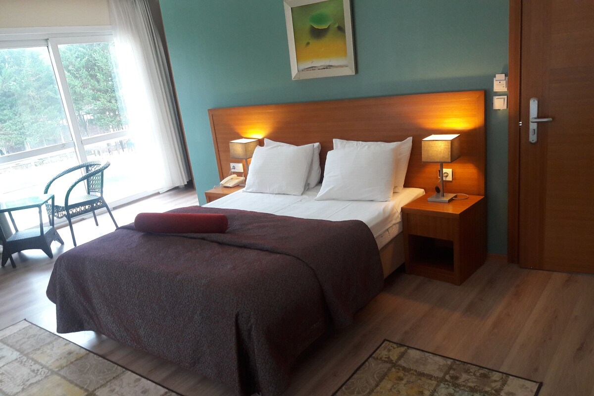 A neatly made bed with white linens and a brown bedspread, two bedside lamps, a wooden headboard, a small bedside table with a lamp, a chair, and a window with curtains.