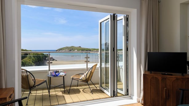 A balcony with a view of the beach, a television on a wooden cabinet, and two chairs with a table. - The Beach House & Porth  Sands Apartments (Newquay)