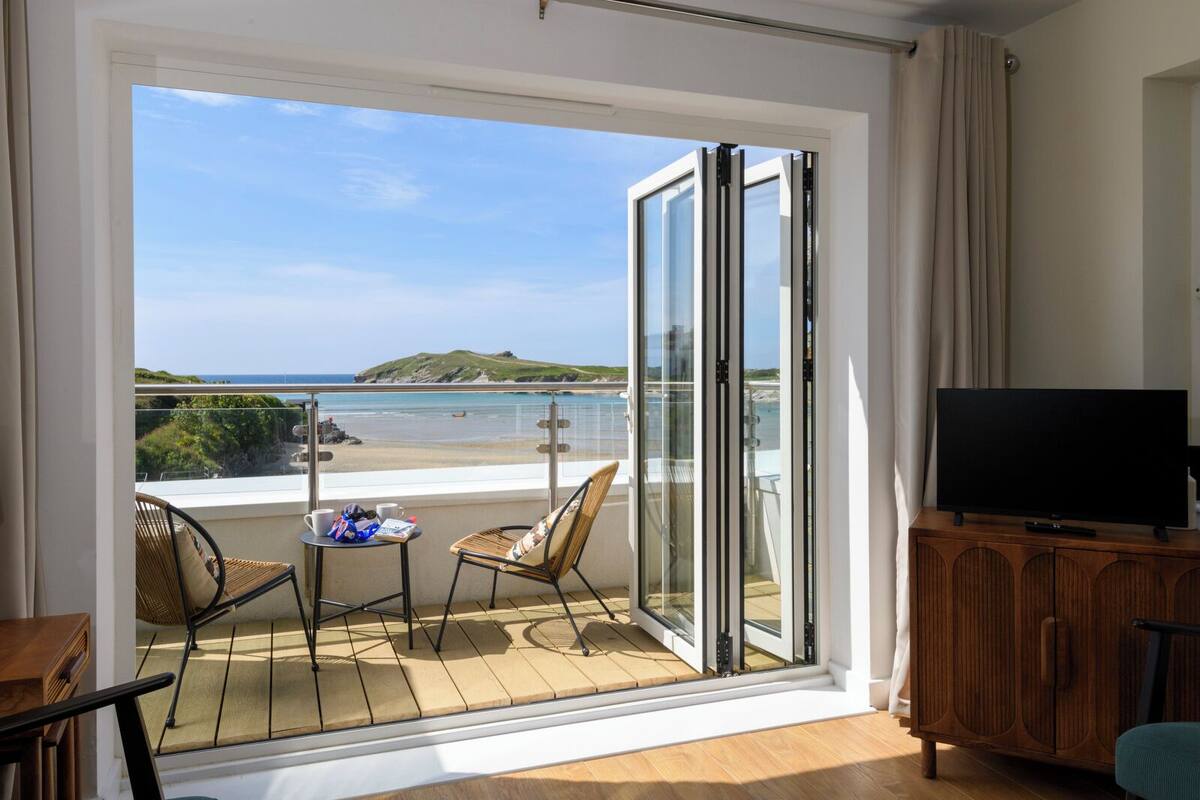 Un balcon donnant sur la plage, un téléviseur posé sur un meuble en bois, et deux chaises avec une table.