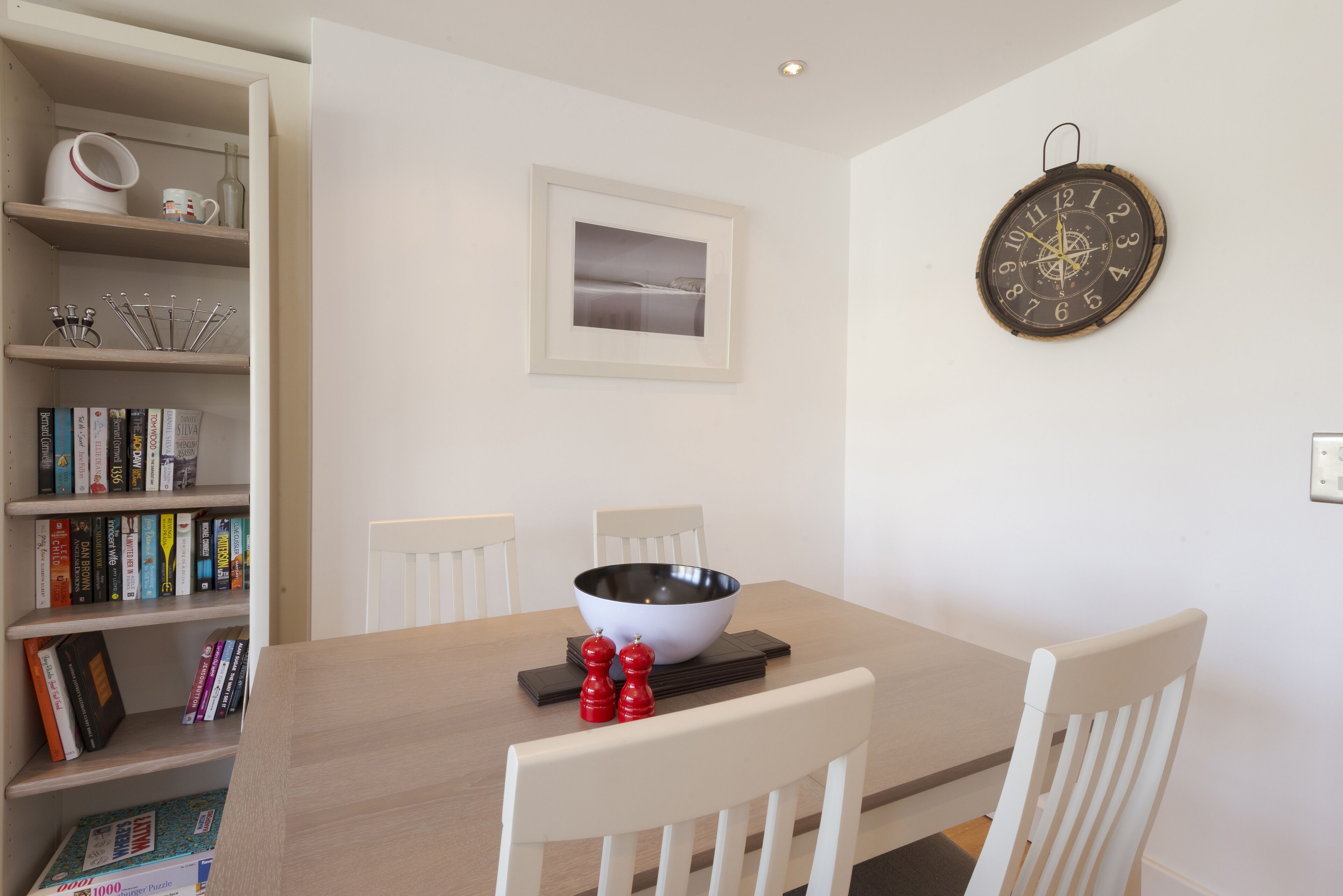 Un área de comedor con una mesa de madera, sillas blancas, un tazón, saleros y pimenteros, una estantería con libros y un reloj en la pared.
