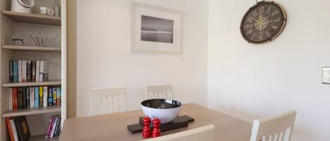 Un área de comedor con una mesa de madera, sillas blancas, un tazón, saleros y pimenteros, una estantería con libros y un reloj en la pared.