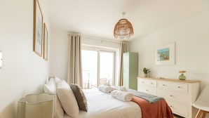 A bedroom with a bed, bedside table, lamp, dresser, and a window with curtains. - The Beach House & Porth  Sands Apartments (Newquay)