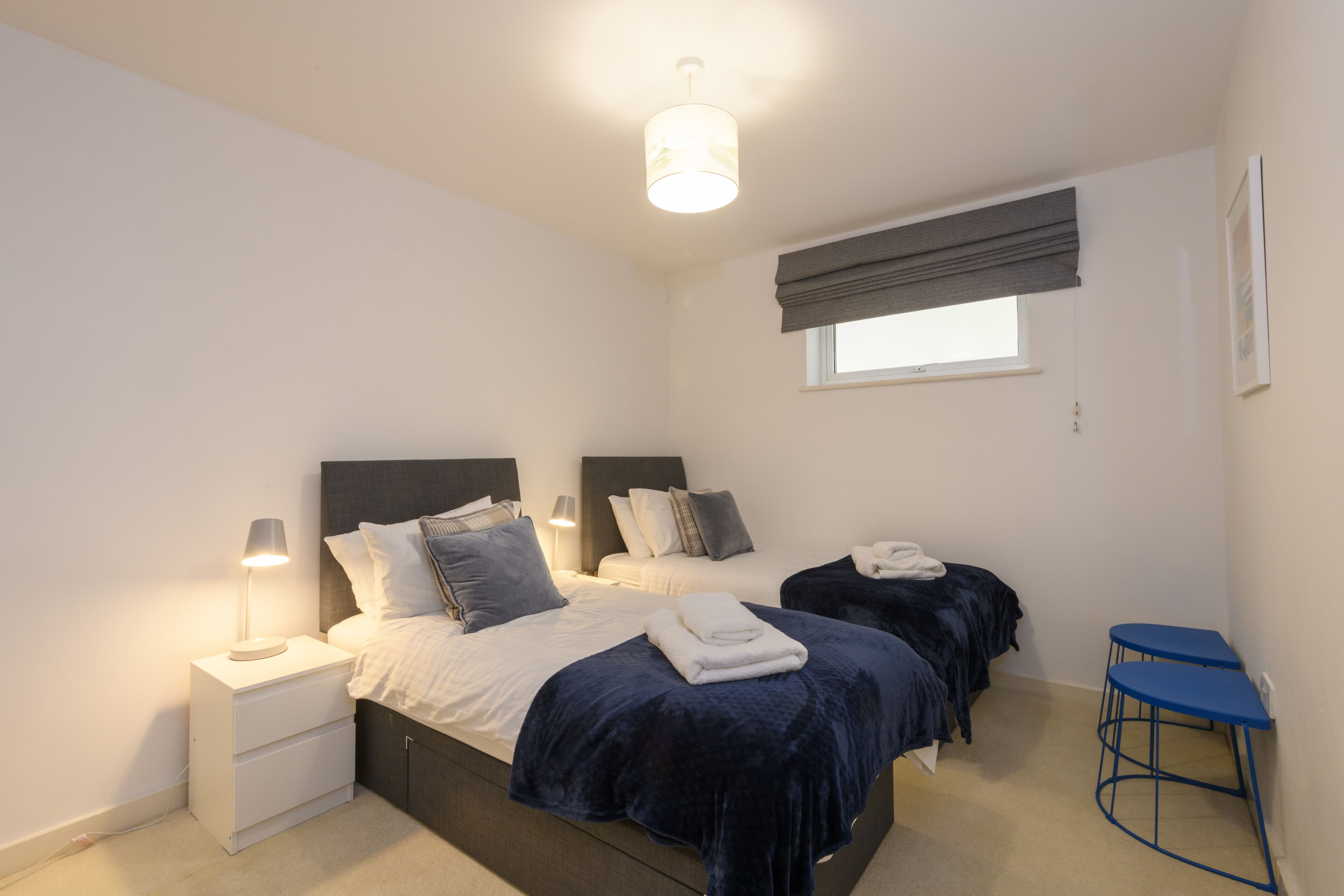 Un dormitorio con dos camas, una mesita de noche con lámpara, una ventana con persiana y una silla azul.