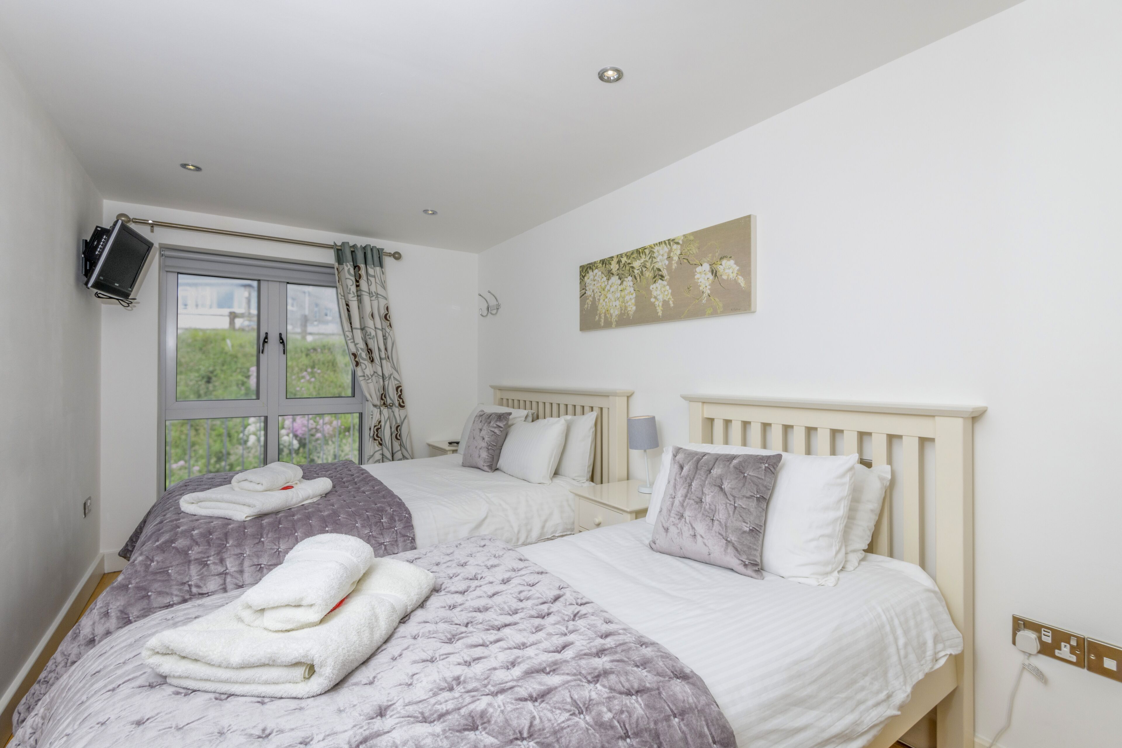 A bedroom with two beds, a window with curtains, a mounted TV, and a wall-mounted artwork.