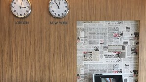 Uma área de recepção com dois relógios identificados com 'LONDON' e 'NEW YORK', um computador de mesa e um vaso de planta.