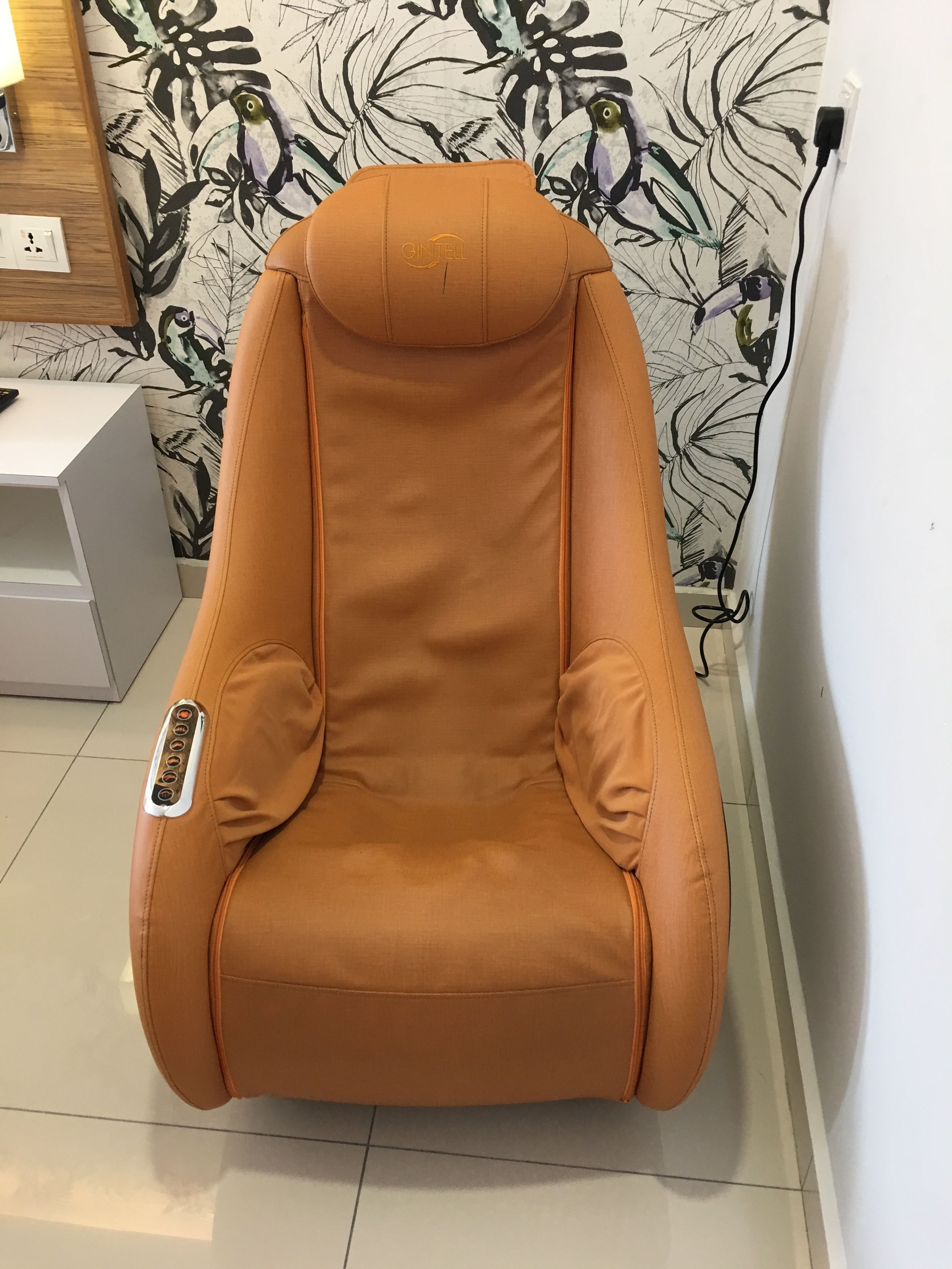 Una cadira de massatge color bronze amb un panell de control, col·locada davant d'una paret amb paper tapet floral.