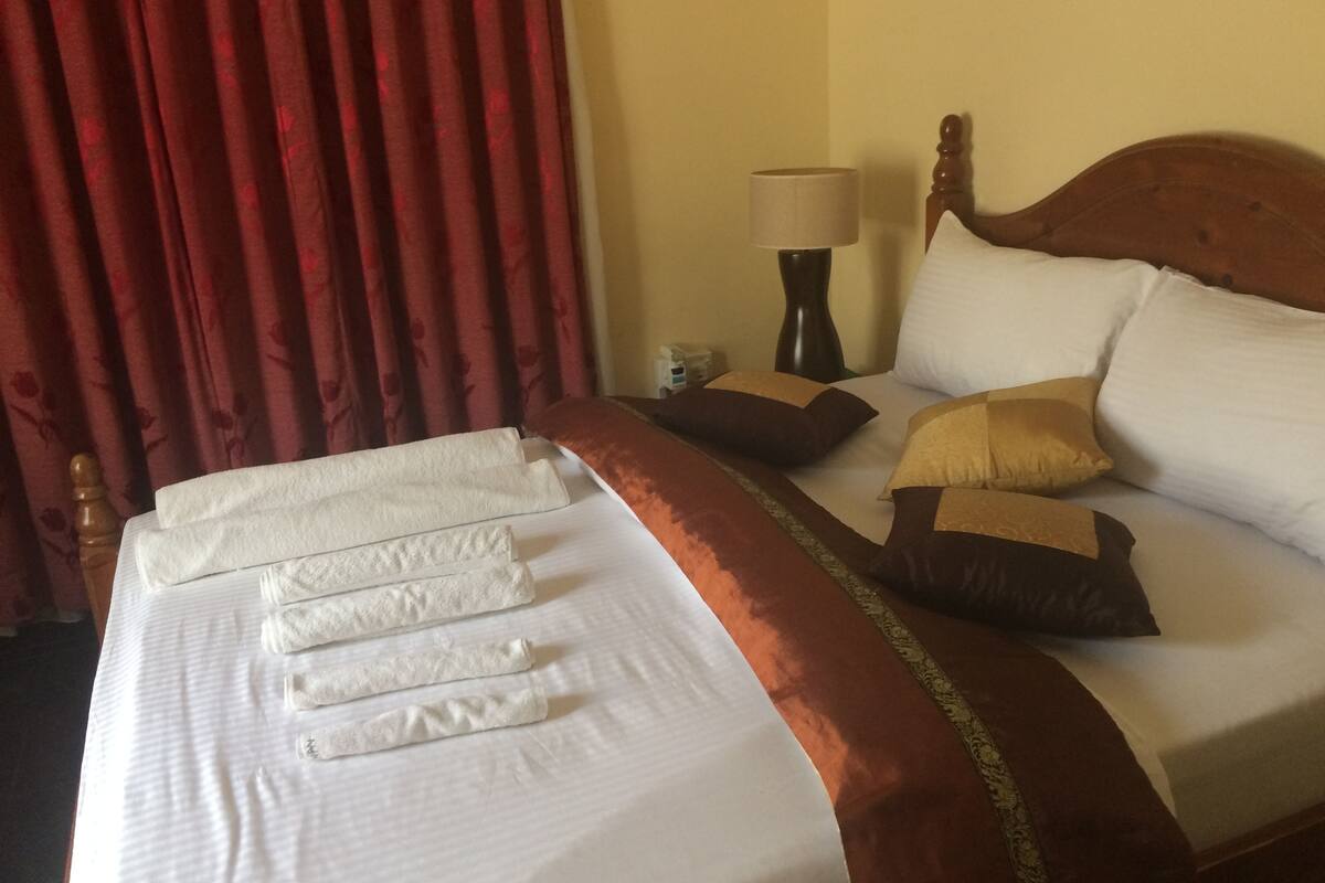 A neatly made bed with white linens and a brown headboard, a lamp on the nightstand, and red curtains in the background.