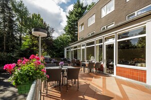 Ein Terrassenbereich im Freien mit Tischen und Stühlen, umgeben von einem Ziegelgebäude mit großen Fenstern.