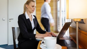 A woman using a laptop at a table with a white cup. There is a door and a window in the background. - Messehotel Köln-Deutz (Cologne)