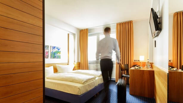 A hotel room with a bed, a desk, a chair, and a TV mounted on the wall. - Messehotel Köln-Deutz (Cologne)